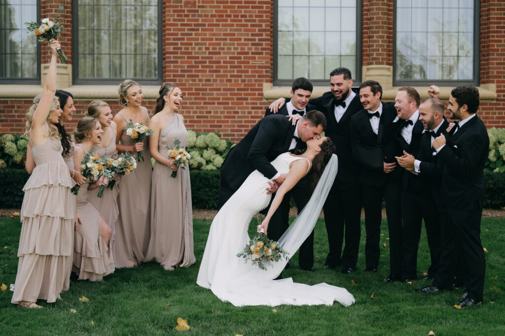 Bride and groom kiss, surrounded by wedding party.